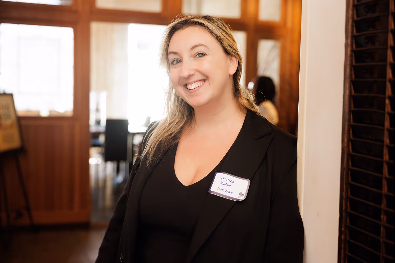 Smiling woman with blonde hair leaning against a wall, wearing black and a name tag.