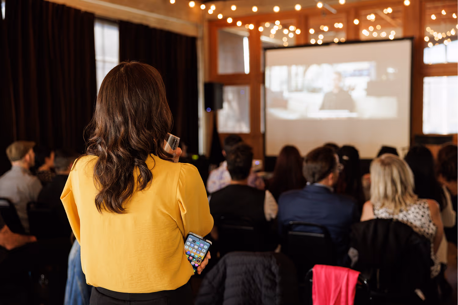 Woman in yellow shirt holding a smartphone, facing an audience watching a presentation on a screen.