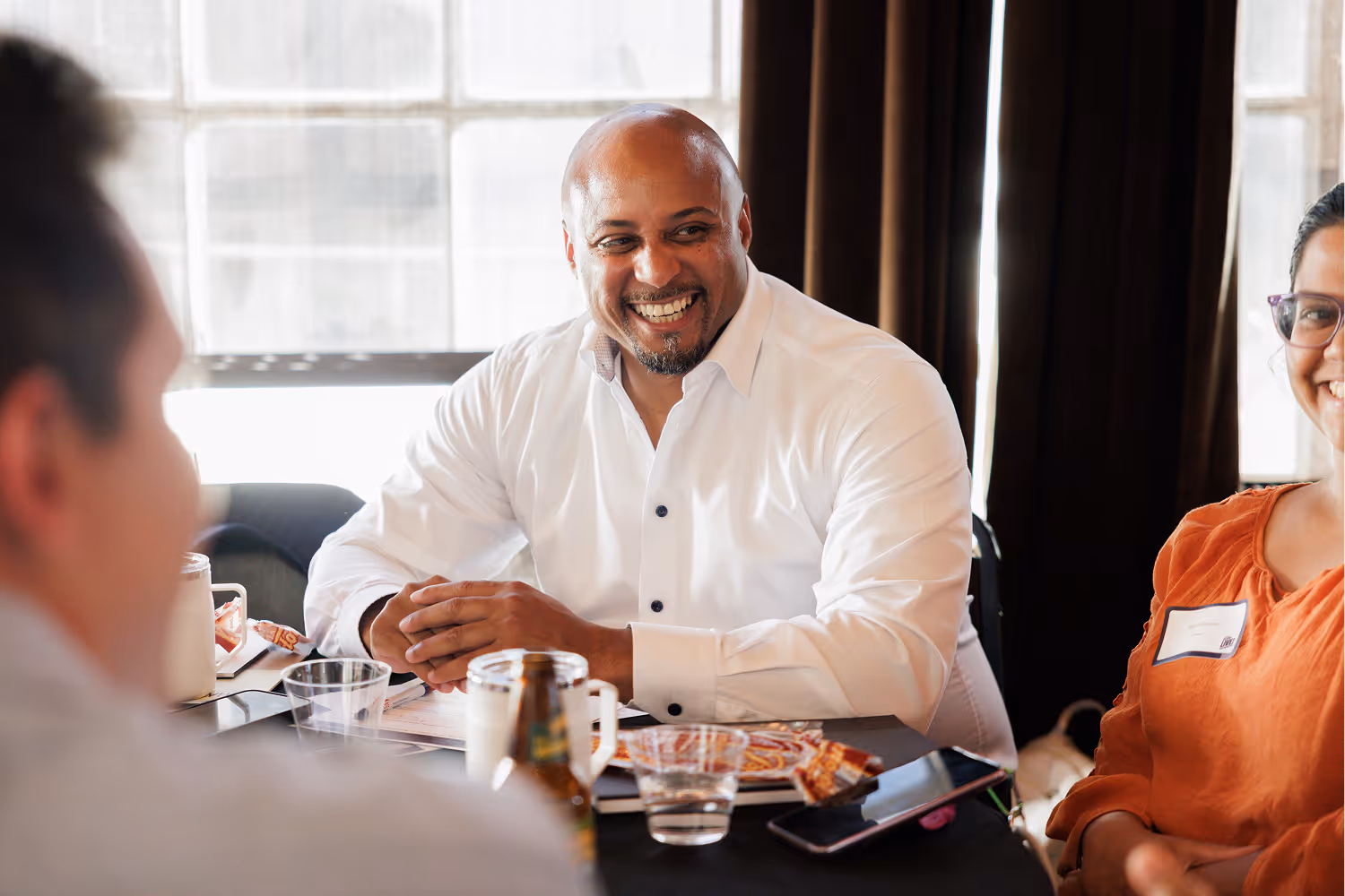 Smiling man in white shirt sitting at table with people, drinks, and snacks in a bright room.