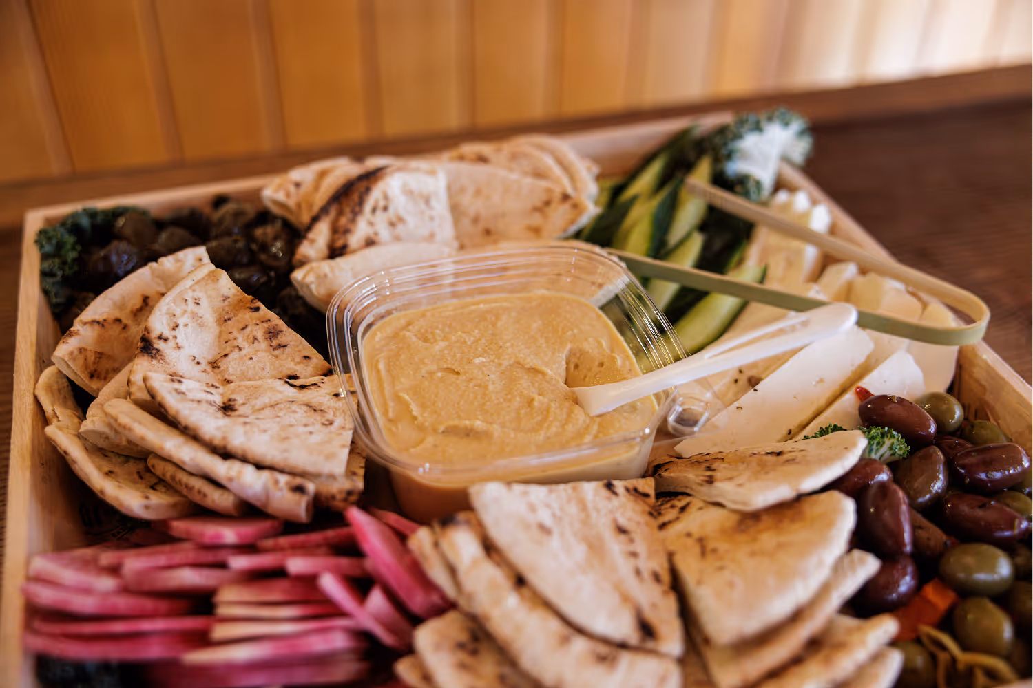 Tray with pita bread, hummus, olives, cucumber, cheese, and sliced radishes on a wooden table.