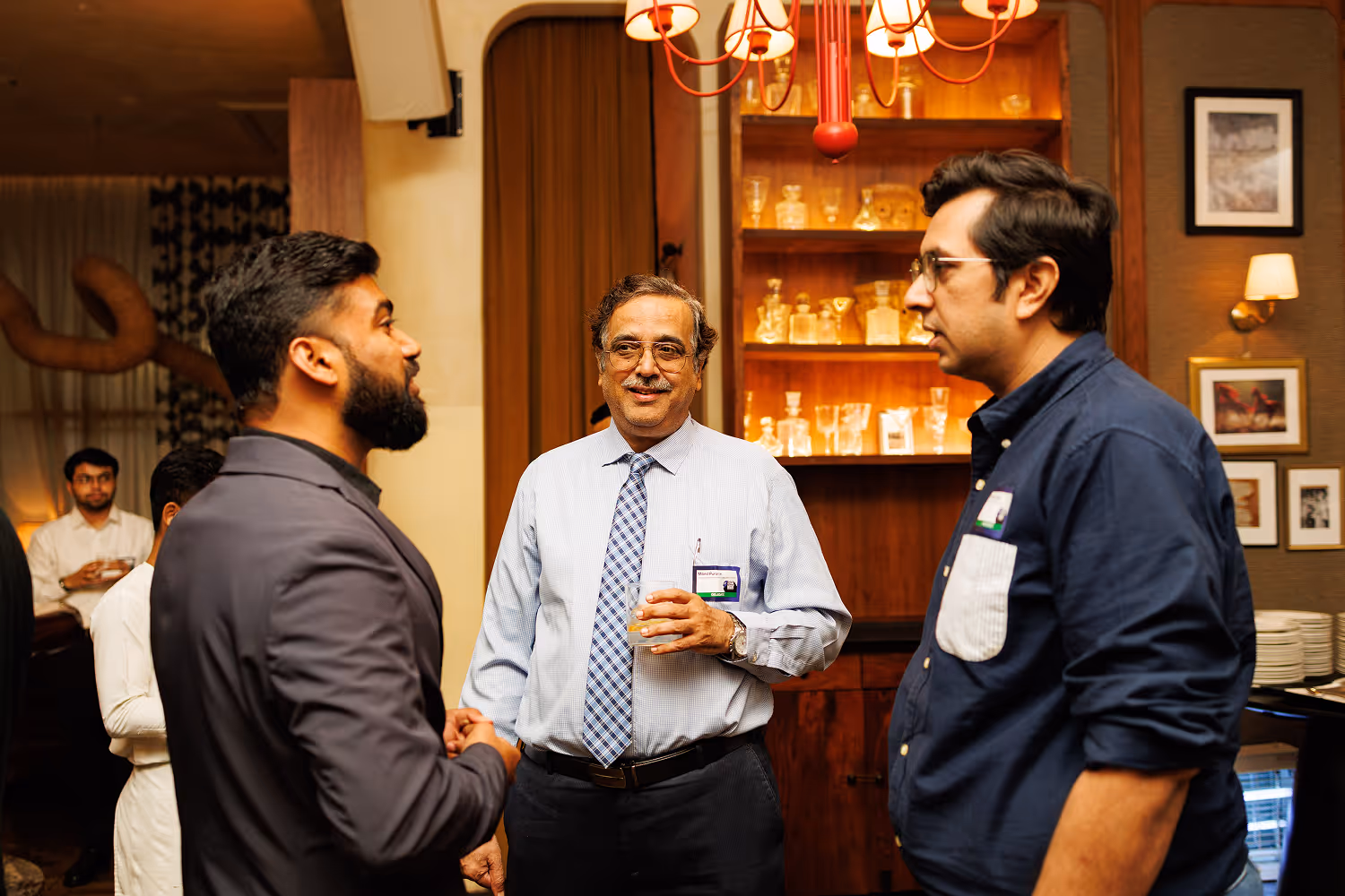 Three men in conversation at a social event in a warmly lit room with shelves and framed pictures.