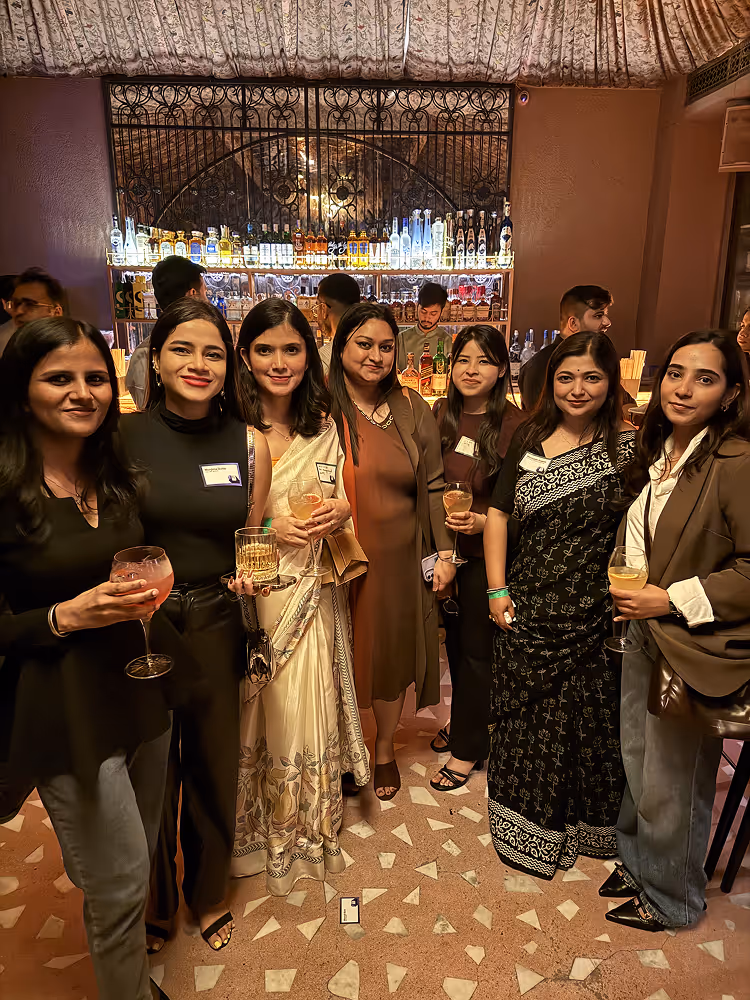 Seven women posing with drinks in a warmly lit bar with bottles displayed behind them.