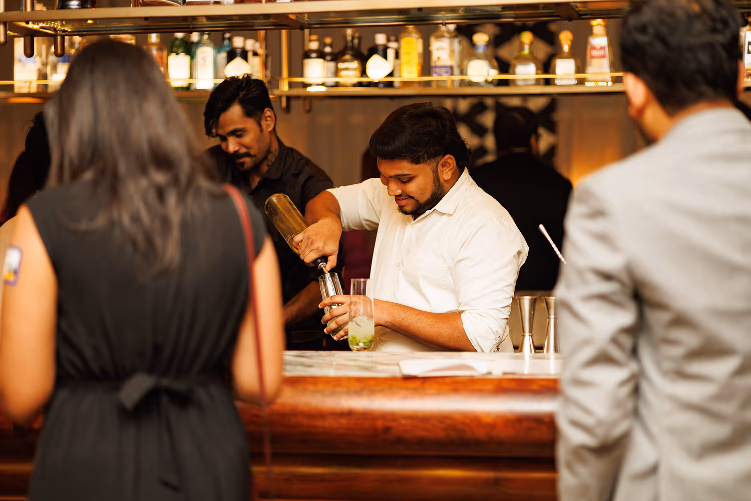 Bartender in white shirt pouring a drink behind a wooden bar with customers in foreground.