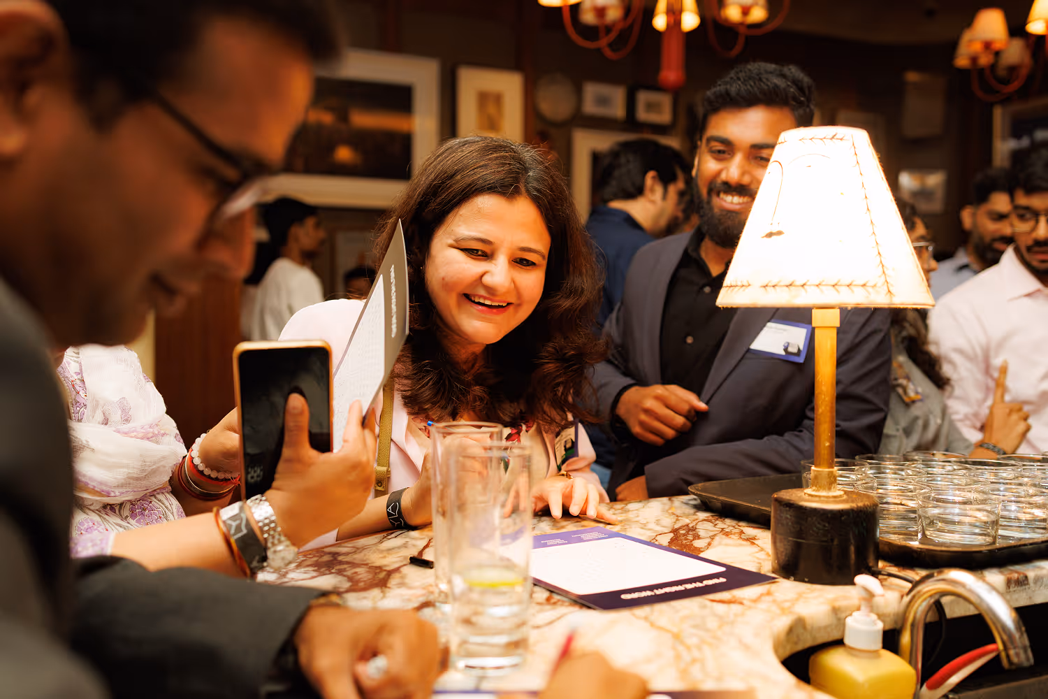 People smiling and interacting at a marble countertop with a small lamp and tray of glasses.