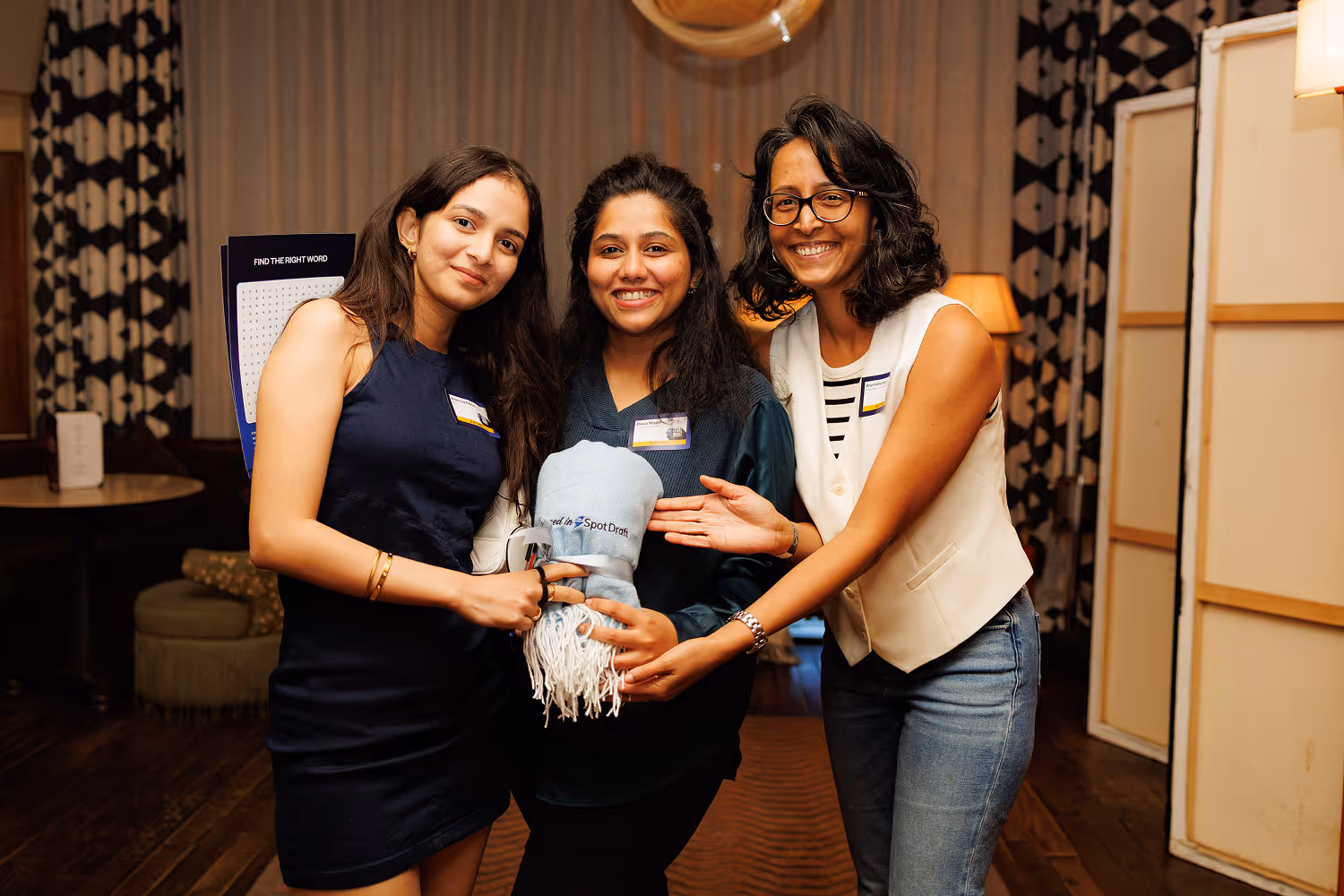 Three smiling women standing together, holding a blue scarf with a logo, indoors with patterned curtains.