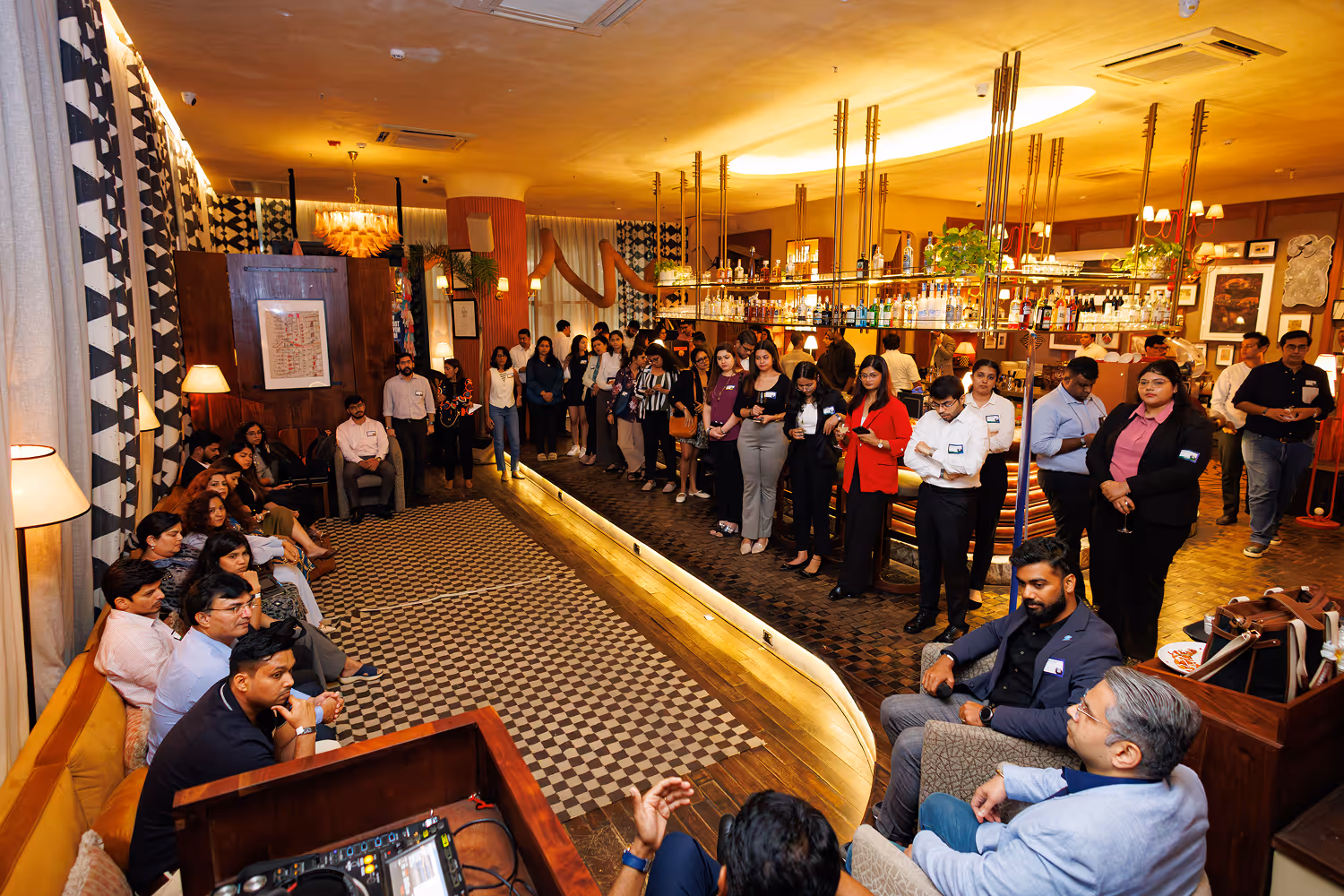 Group of people standing and sitting inside a warmly lit bar or lounge with patterned carpet.
