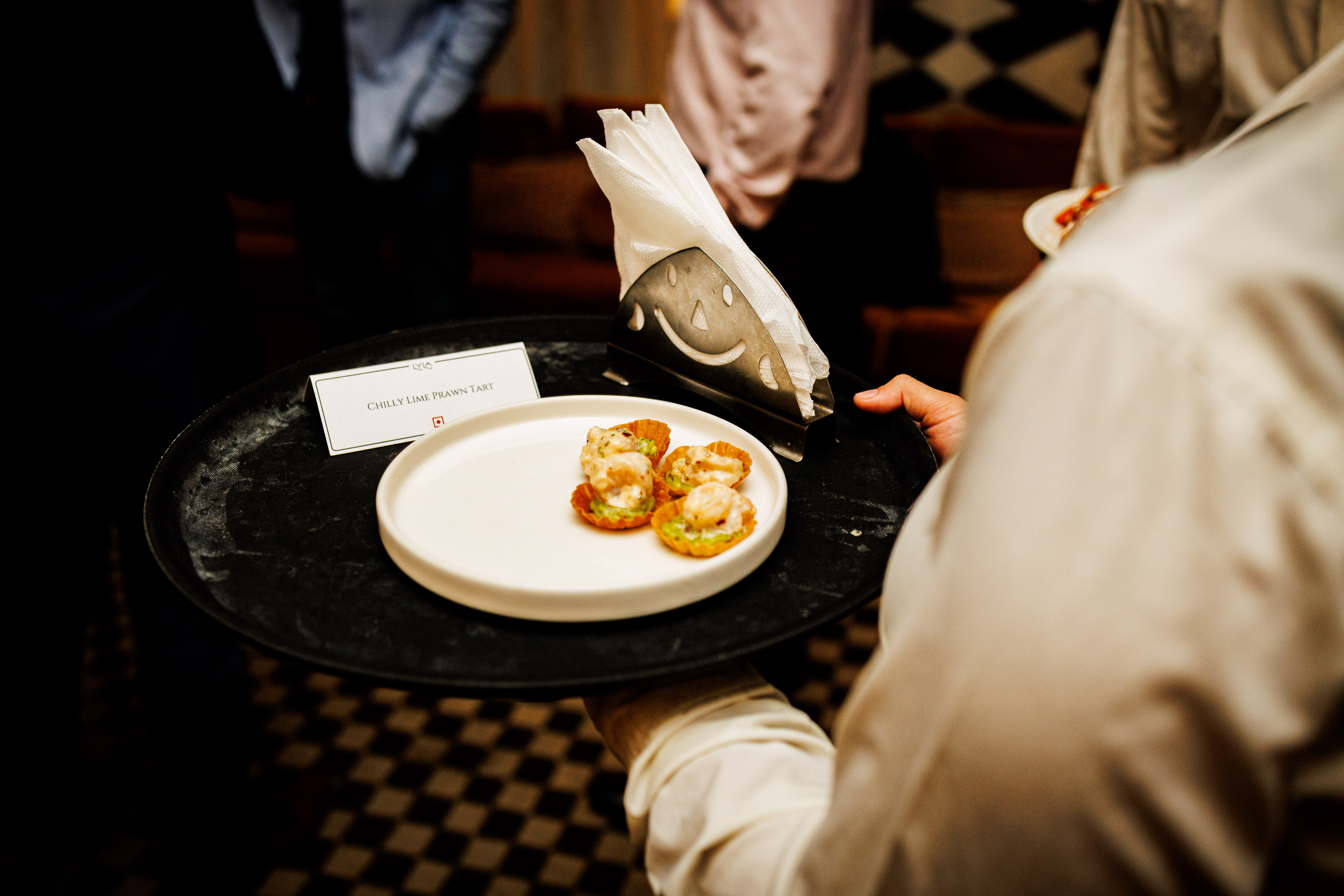 Server holding tray with plate of four chilly lime prawn tarts and a napkin holder.