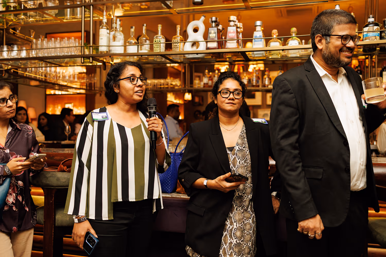 Group of people in a bar setting; one woman speaks into a microphone while others listen and hold phones.