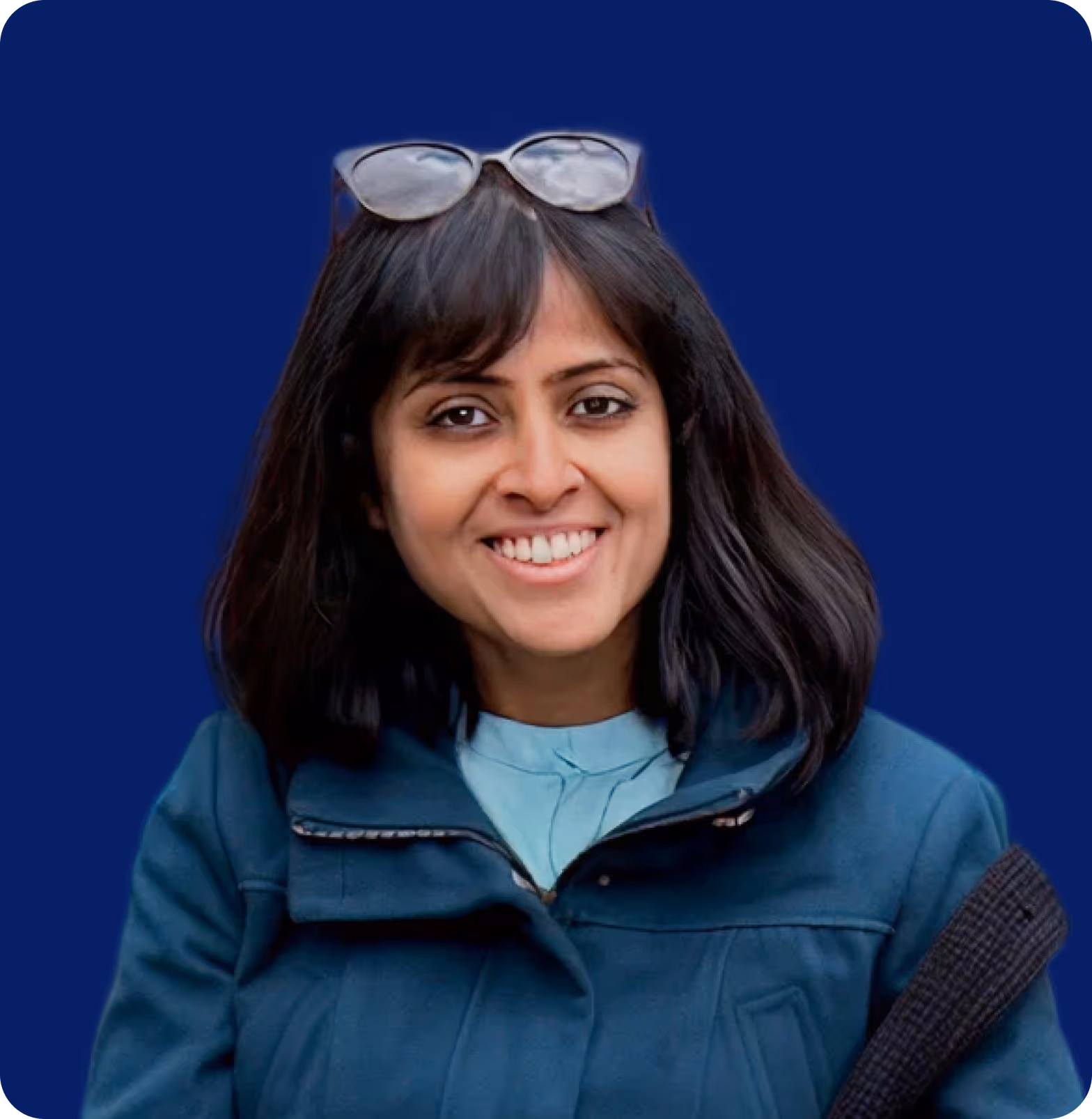 Smiling woman with dark hair wearing glasses on her head and a blue jacket against a blue background.