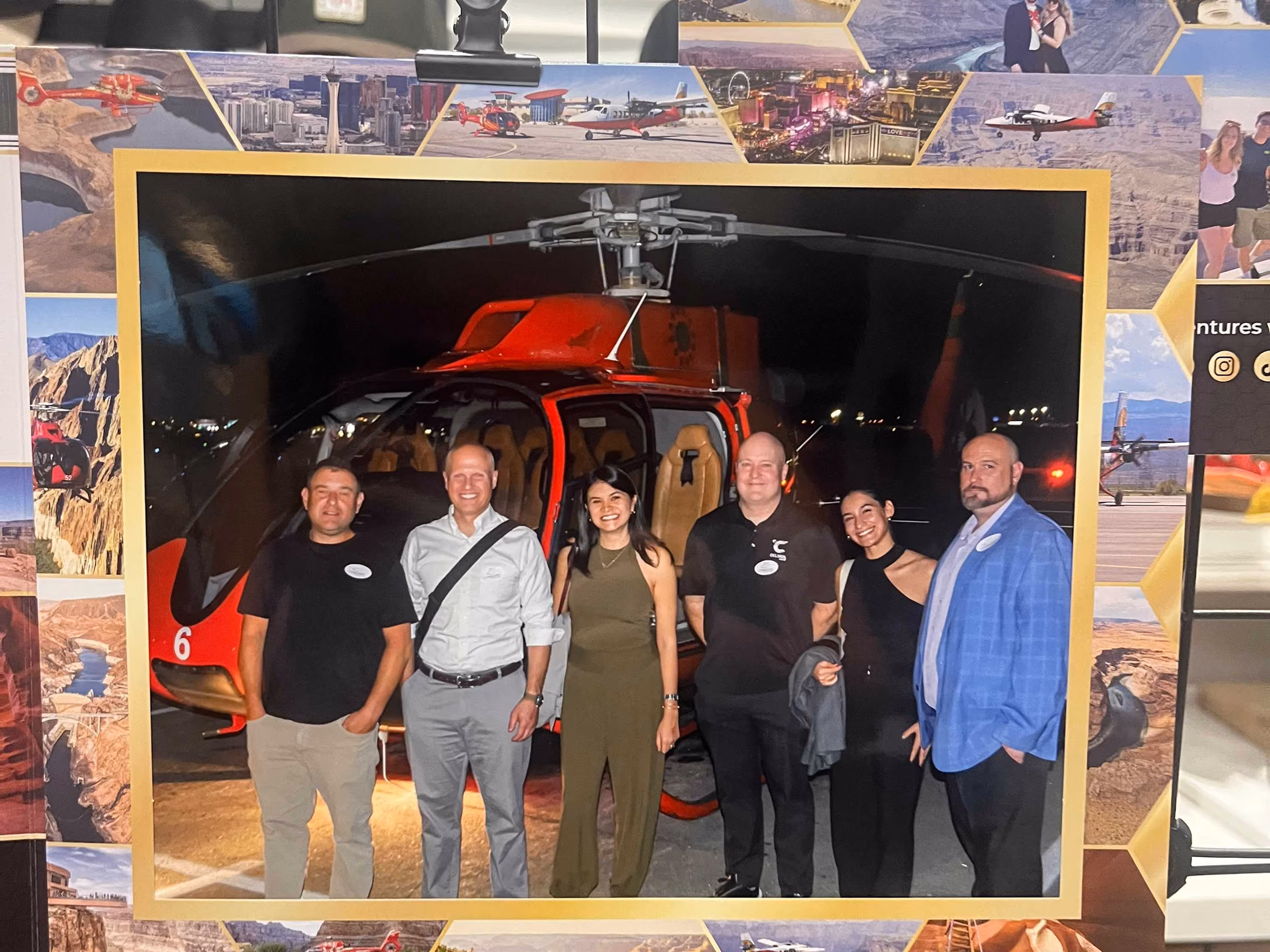 Five adults standing and smiling in front of a red helicopter at night, all posing for the camera in casual and semi-formal attire.