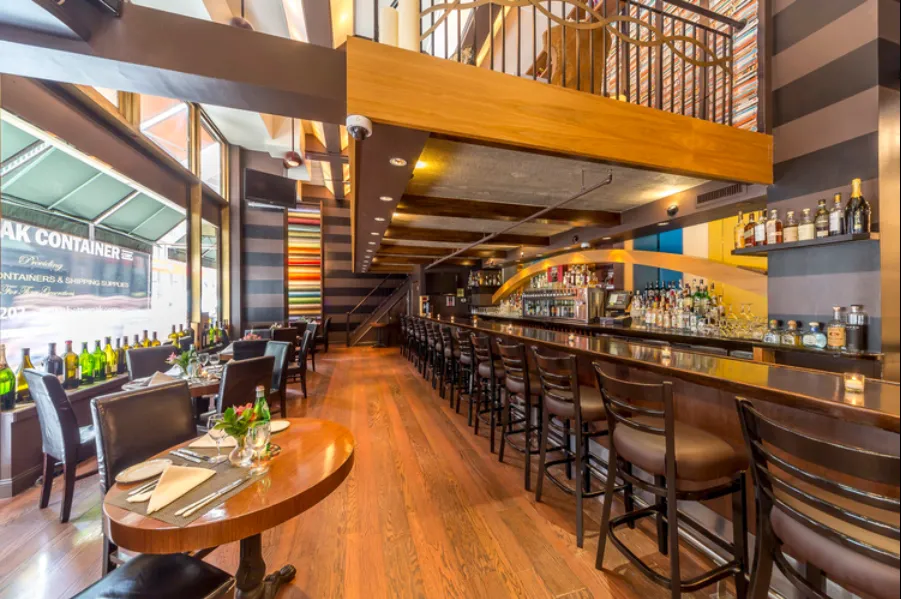 Interior of a modern bar and restaurant with wooden floors, high bar stools along the counter, dining tables by large windows, and colorful wall decor.