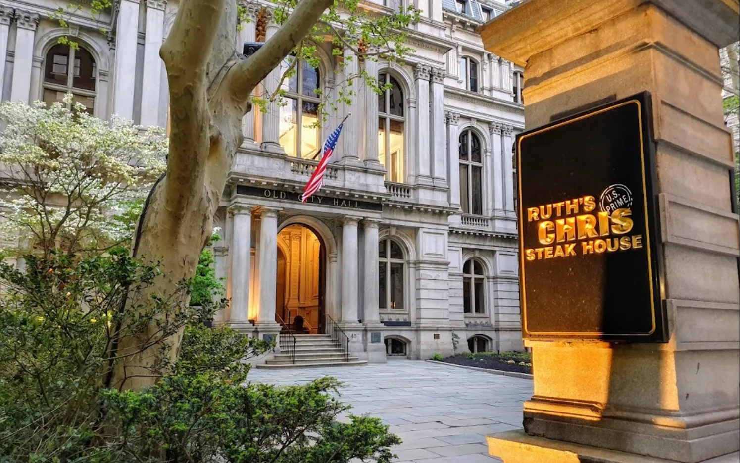 Entrance of Old City Hall building with an American flag and illuminated Ruth's Chris Steak House sign on a stone pillar nearby.