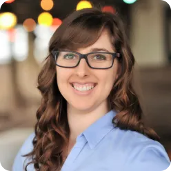 Smiling woman with curly brown hair and glasses wearing a light blue shirt, blurred colorful lights in the background.