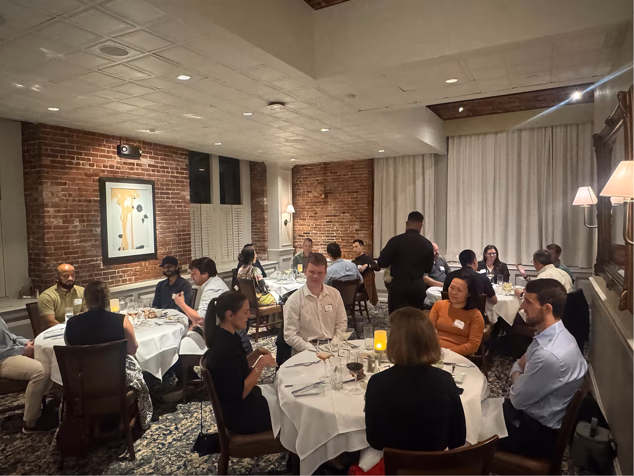 Groups of people sitting at round tables having dinner in a warmly lit restaurant with brick walls.
