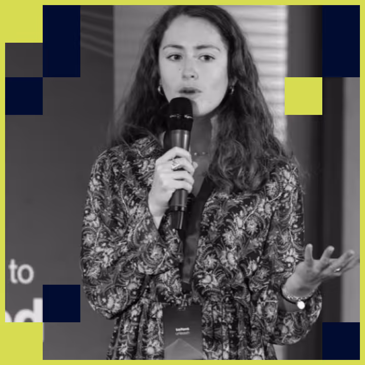 Woman with long curly hair speaking into a microphone, wearing a patterned jacket and gesturing with her hand.