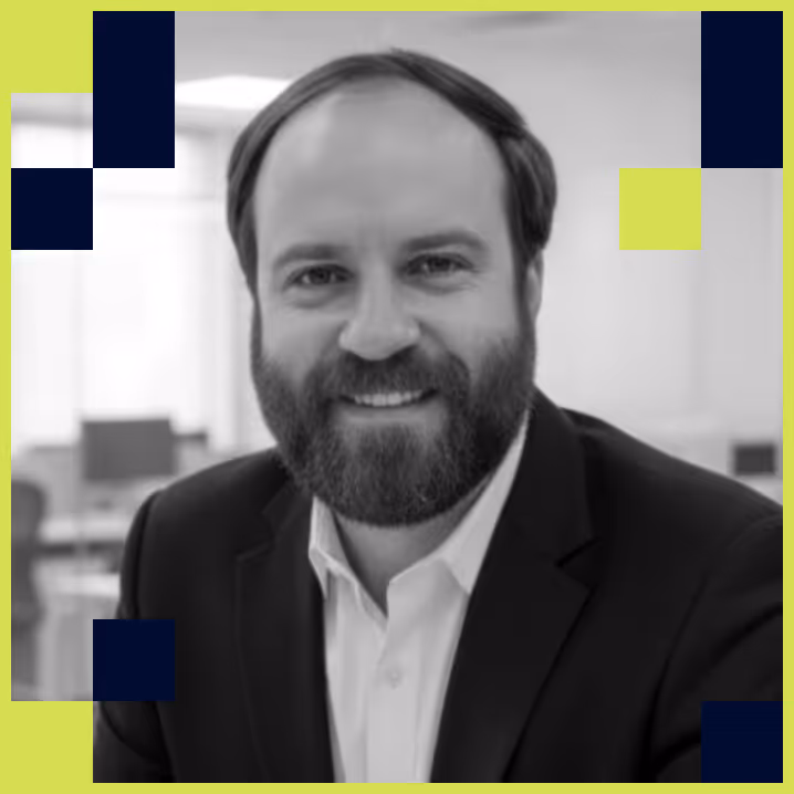 Portrait of a smiling man with a beard wearing a suit jacket and white shirt in an office setting.