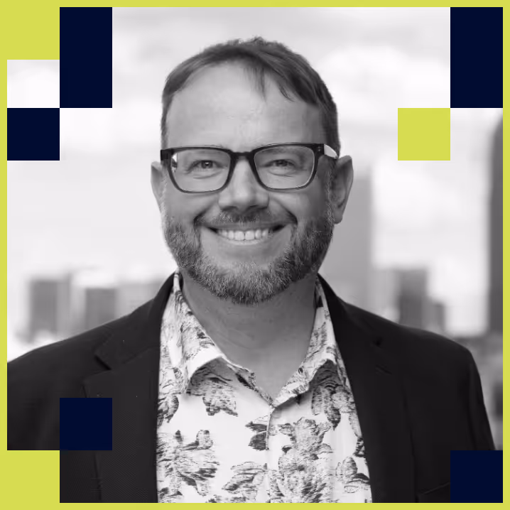 Smiling man with glasses, beard, and floral shirt under a dark blazer, with a blurred cityscape background.