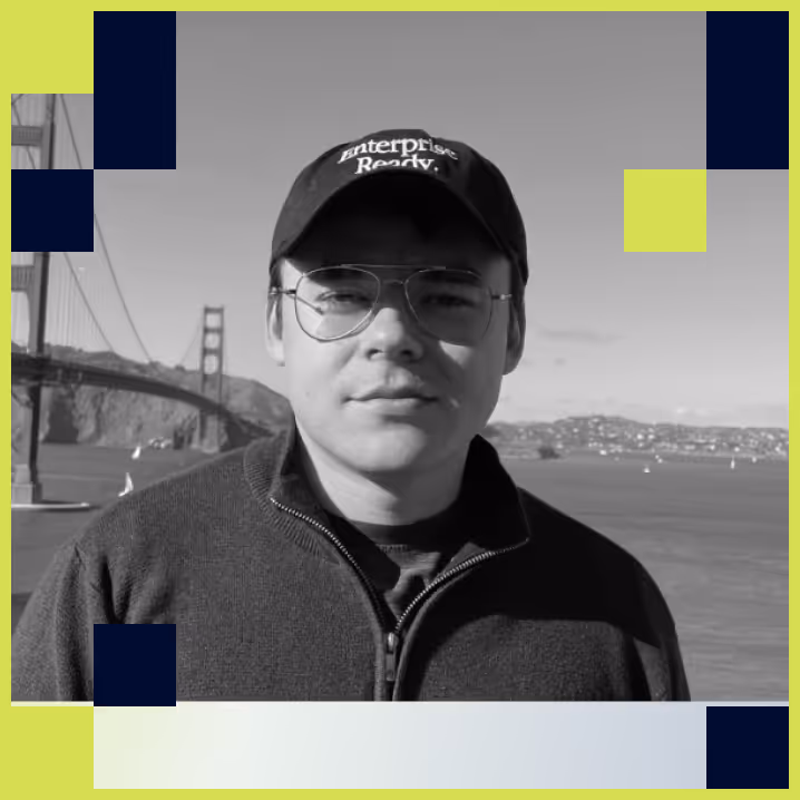 Black-and-white photo of a man wearing glasses and a cap with 'Enterprise Ready' text, standing outdoors with a bridge and water in the background.