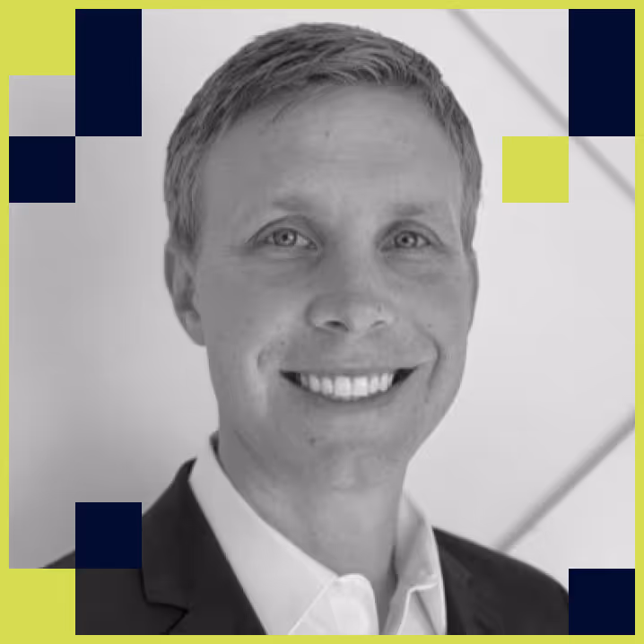Smiling man in a suit jacket and white shirt in a grayscale photo with geometric color accents around the edges.