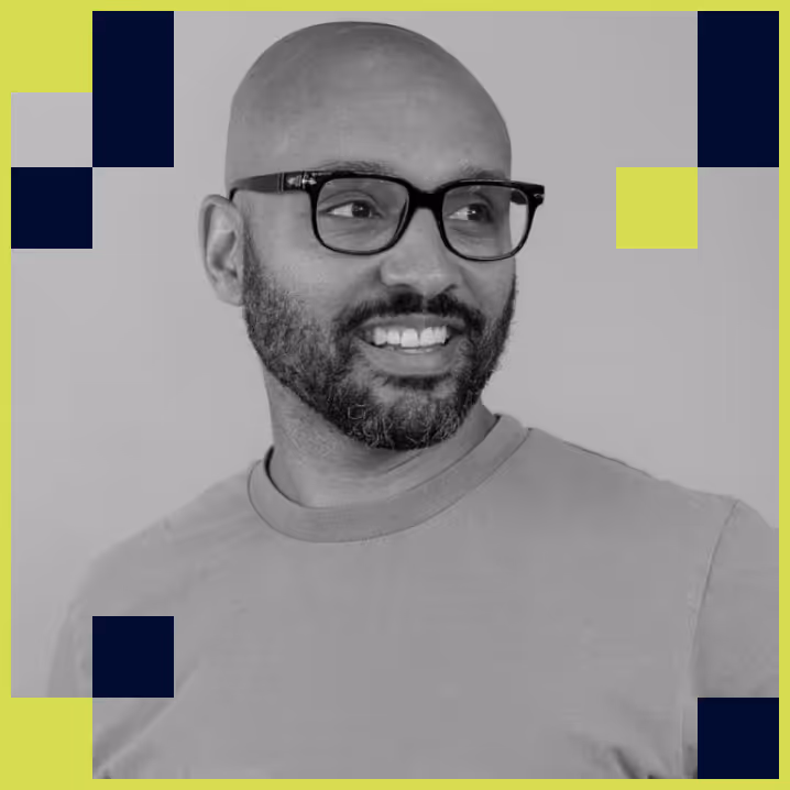 Smiling bald man with a beard and black glasses looking to the side, wearing a light-colored shirt, with decorative navy and lime green squares around the image.