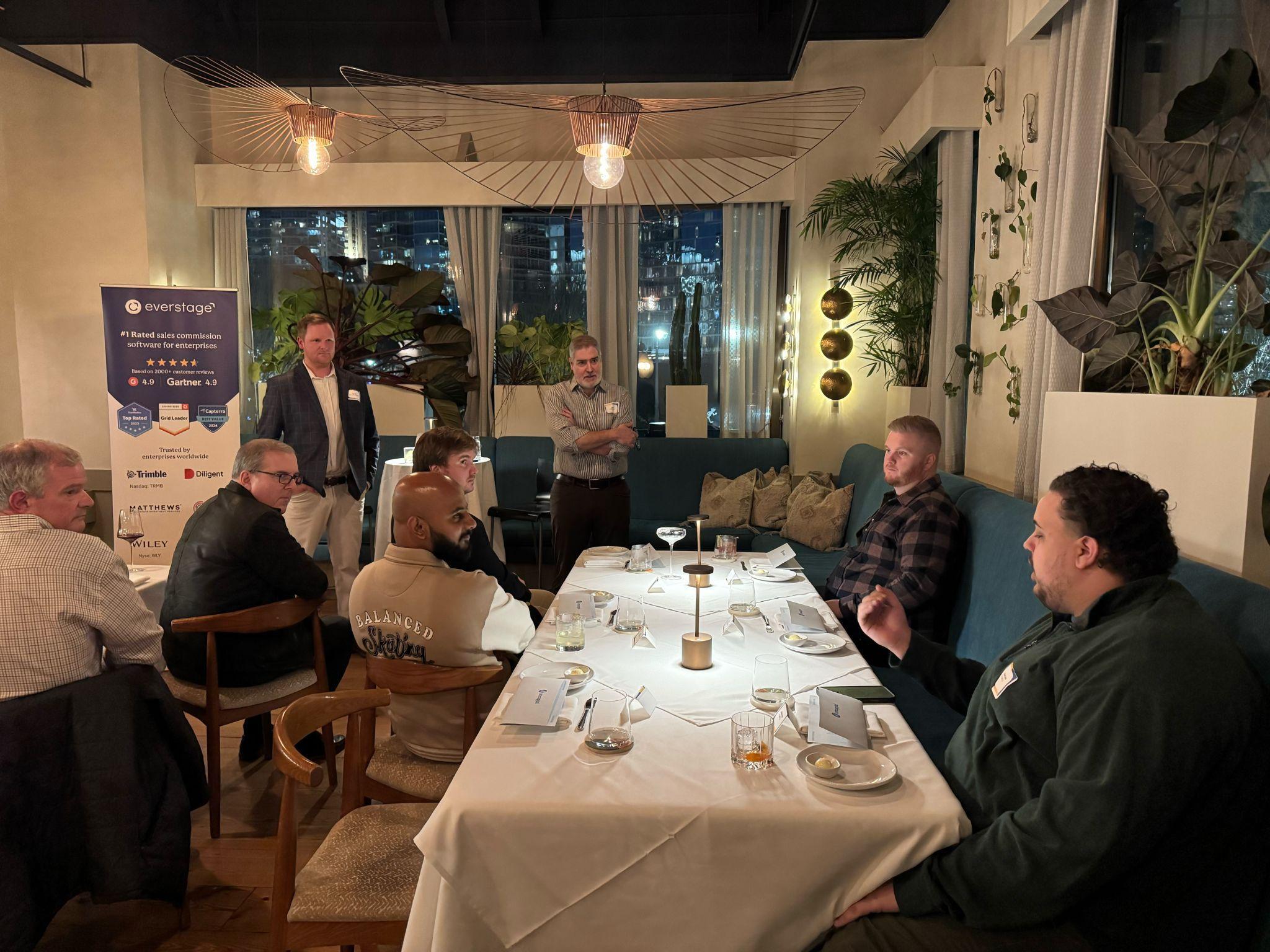 Group of men gathered around a dining table in a modern room with city views, discussing with a standing speaker.