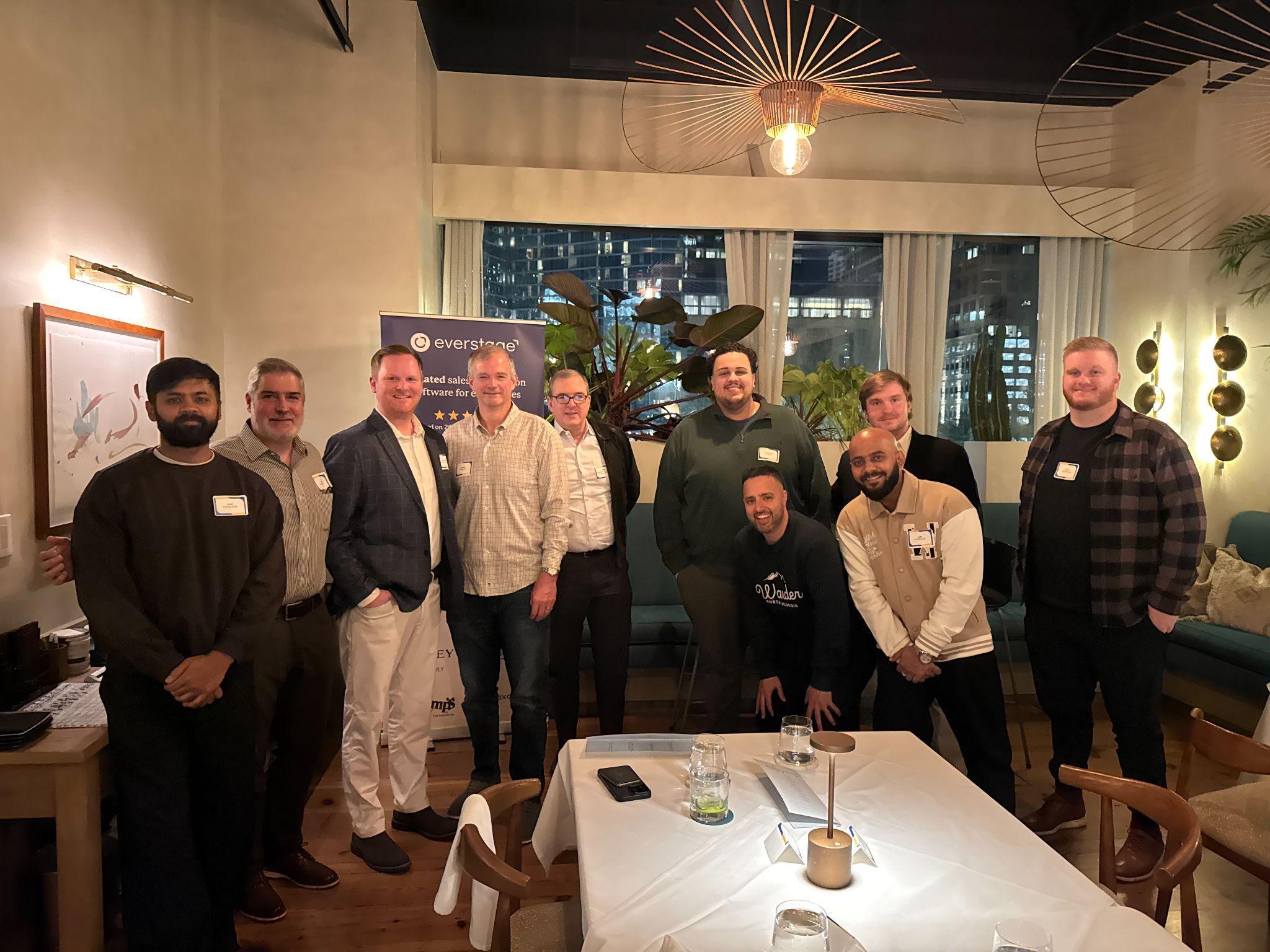 Group of ten men posing indoors in front of a window and a banner under warm lighting in a modern room.