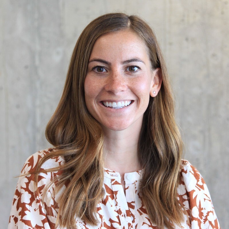Smiling woman with long brown hair wearing floral patterned blouse in front of gray wall.