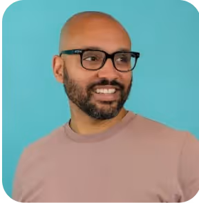 Smiling bald man with glasses and a beard wearing a mauve shirt against a teal background.