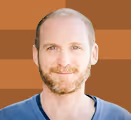 Smiling bald man with light beard wearing a blue shirt against a brown background.