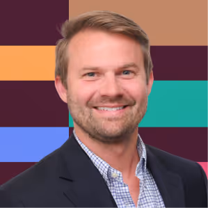 Smiling man with light brown hair, beard, wearing a checkered shirt and dark blazer against colorful background.