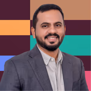 Smiling man with dark hair and beard wearing gray blazer and light shirt in front of colorful background.