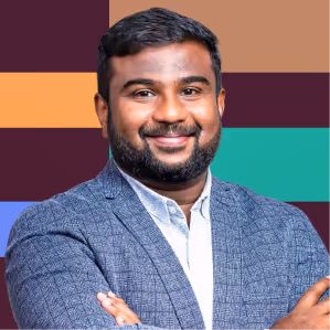 Smiling man with beard in a gray jacket and white shirt against a colorful geometric background.