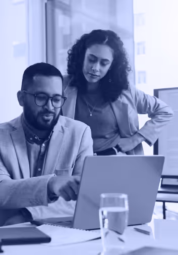 Two coworkers, one sitting and one standing, looking at a laptop in a modern office.