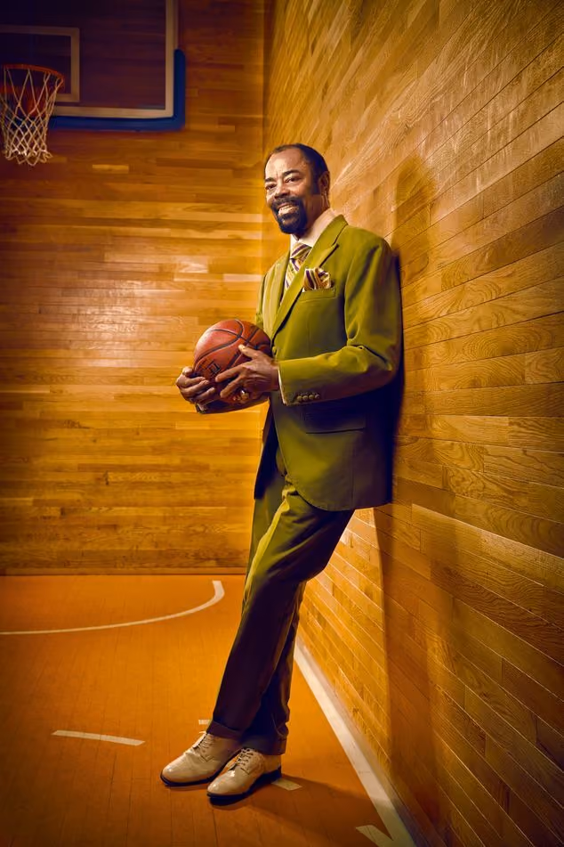 Man in green suit holding basketball and leaning against wooden wall on basketball court.