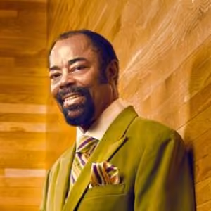 Smiling man with goatee wearing a green suit and striped tie against a wood-paneled background.