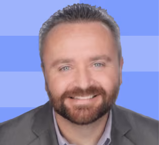 Smiling man with short dark hair and beard wearing gray blazer against blue striped background.