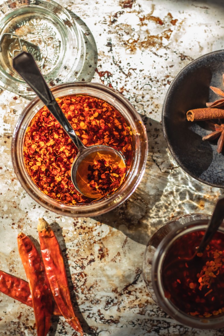 Jar of Sichuan chili oil with a spoon surrounded by dried red chilies and a bowl with cinnamon and star anise