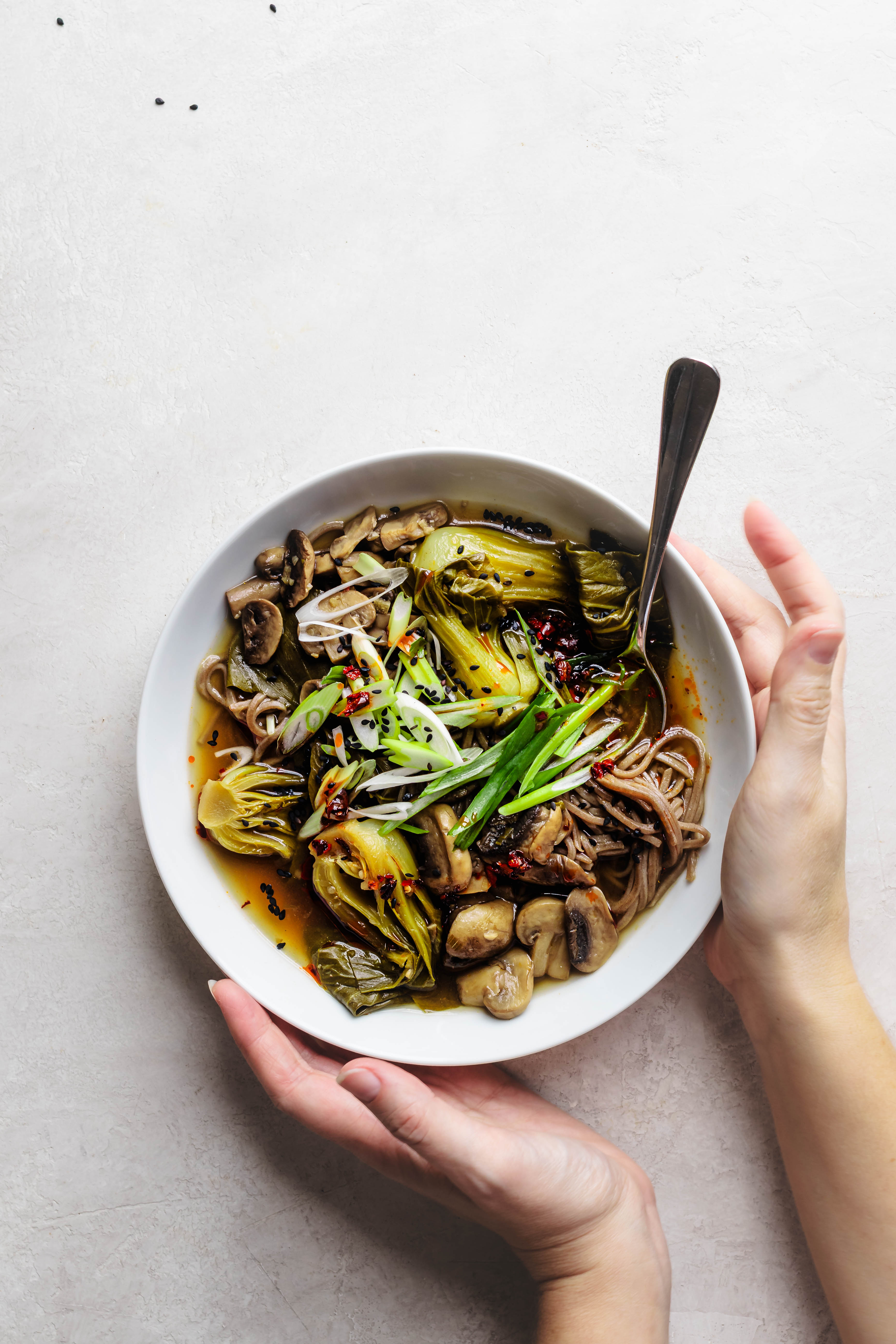 Hands holding a bowl of bok choy mushroom noodle soup with a spoon on a white background