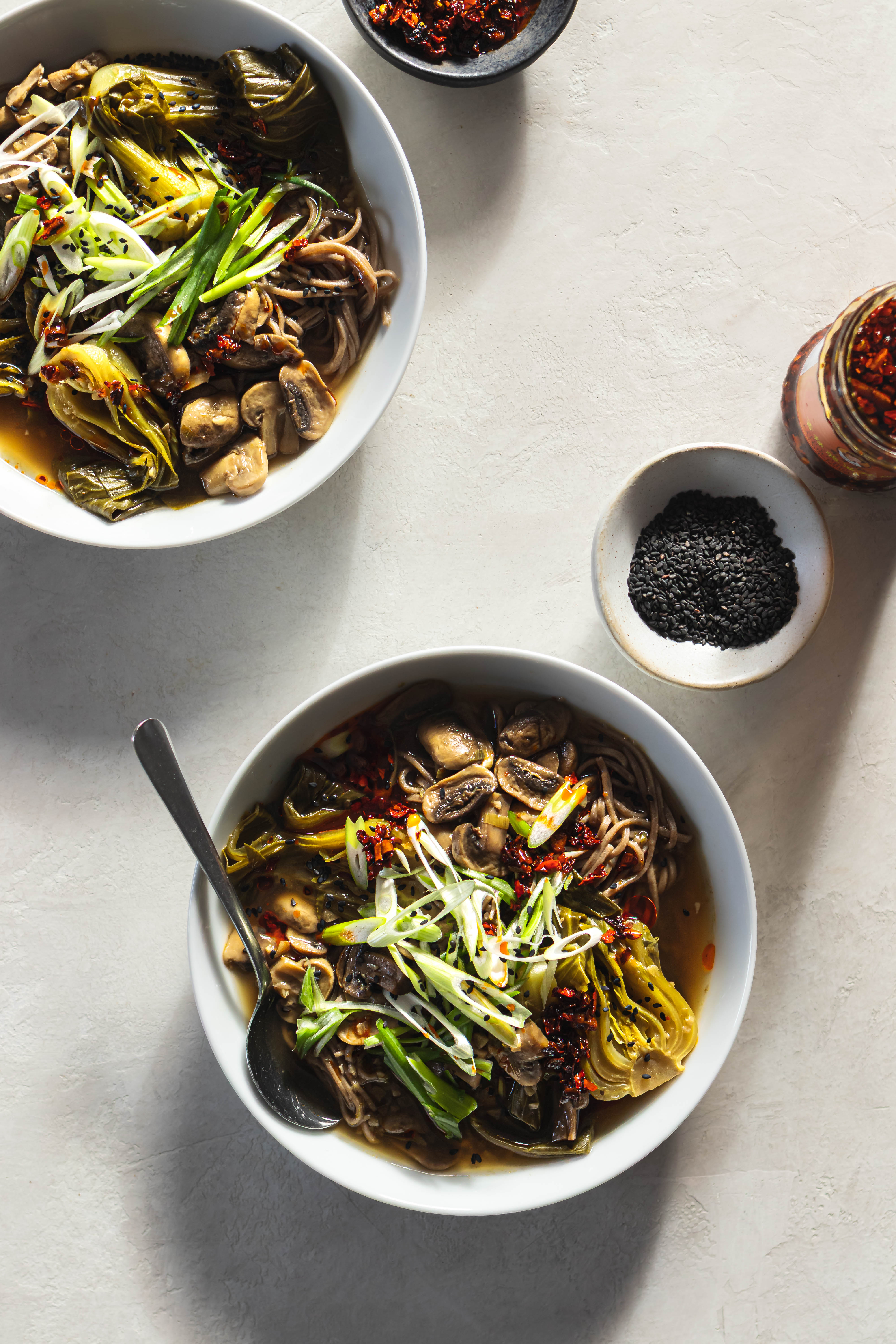 Two bowls of bok choy mushroom noodle soup surrounded by pinch bowls of chili oil and black sesame seeds