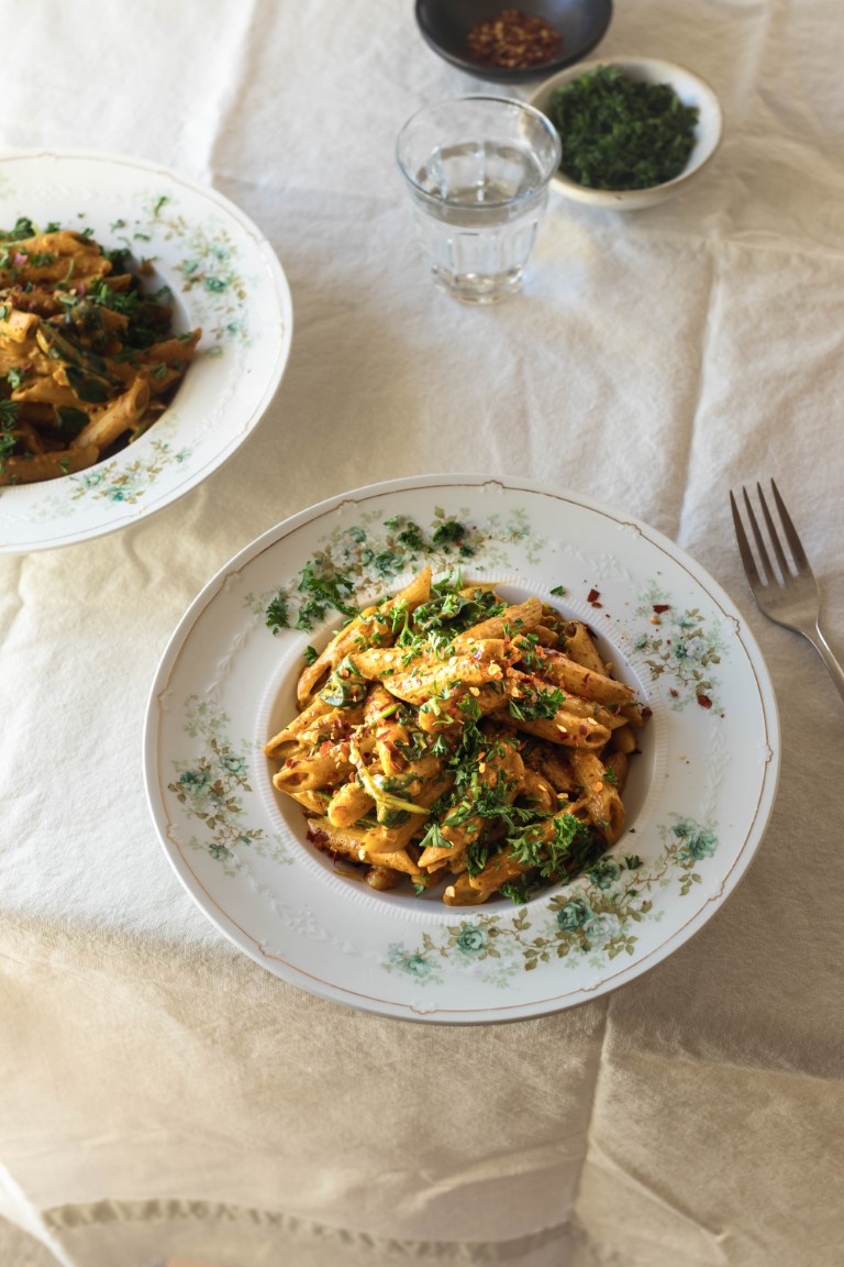 A table covered in a white tablecloth with two antique green floral bowls filled with penne pasta in a creamy curry sauce and a few pinch bowls with chopped herbs and red chili flakes