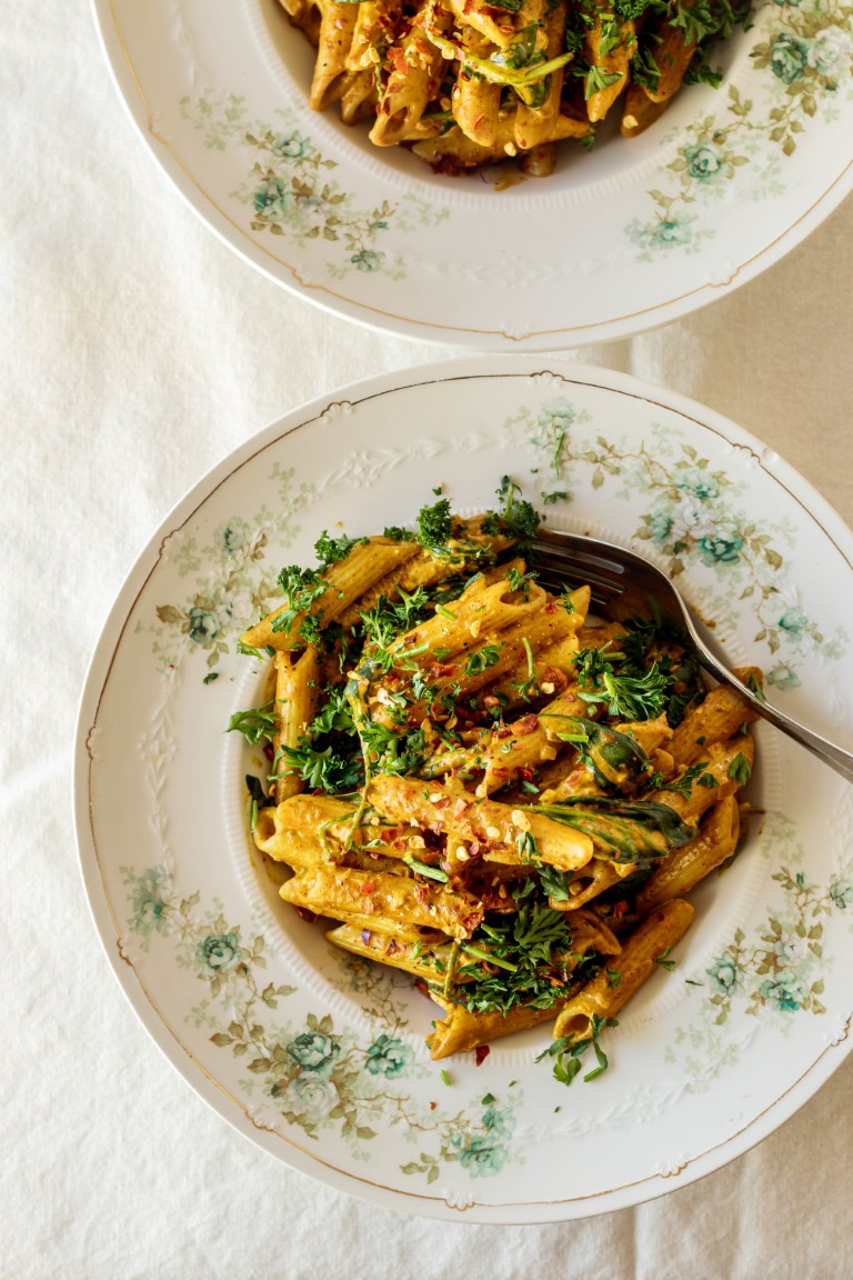 Two large china bowls decorated with a green rose floral pattern sitting on a white tablecloth and filled with a creamy curry penne topped with herbs and red chili flakes