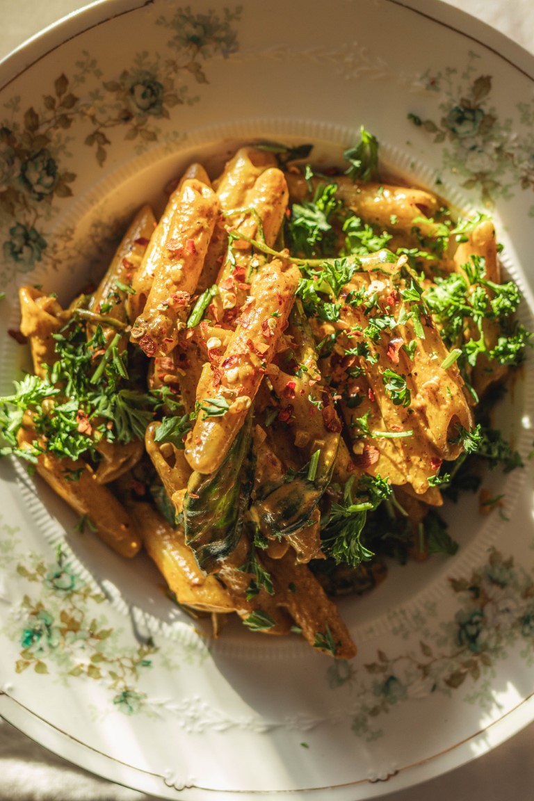 A close up of a bowl of penne in a creamy orange curry sauce garnished with herbs and red chili flakes in direct sunlight