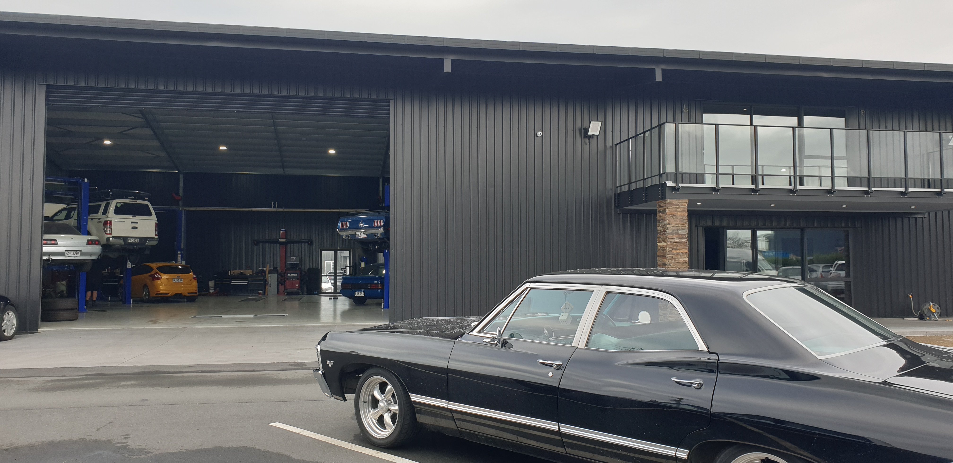 Commercial workshop building in dark steel cladding, with an open bay showing vehicles on hoists and a glazed mezzanine office