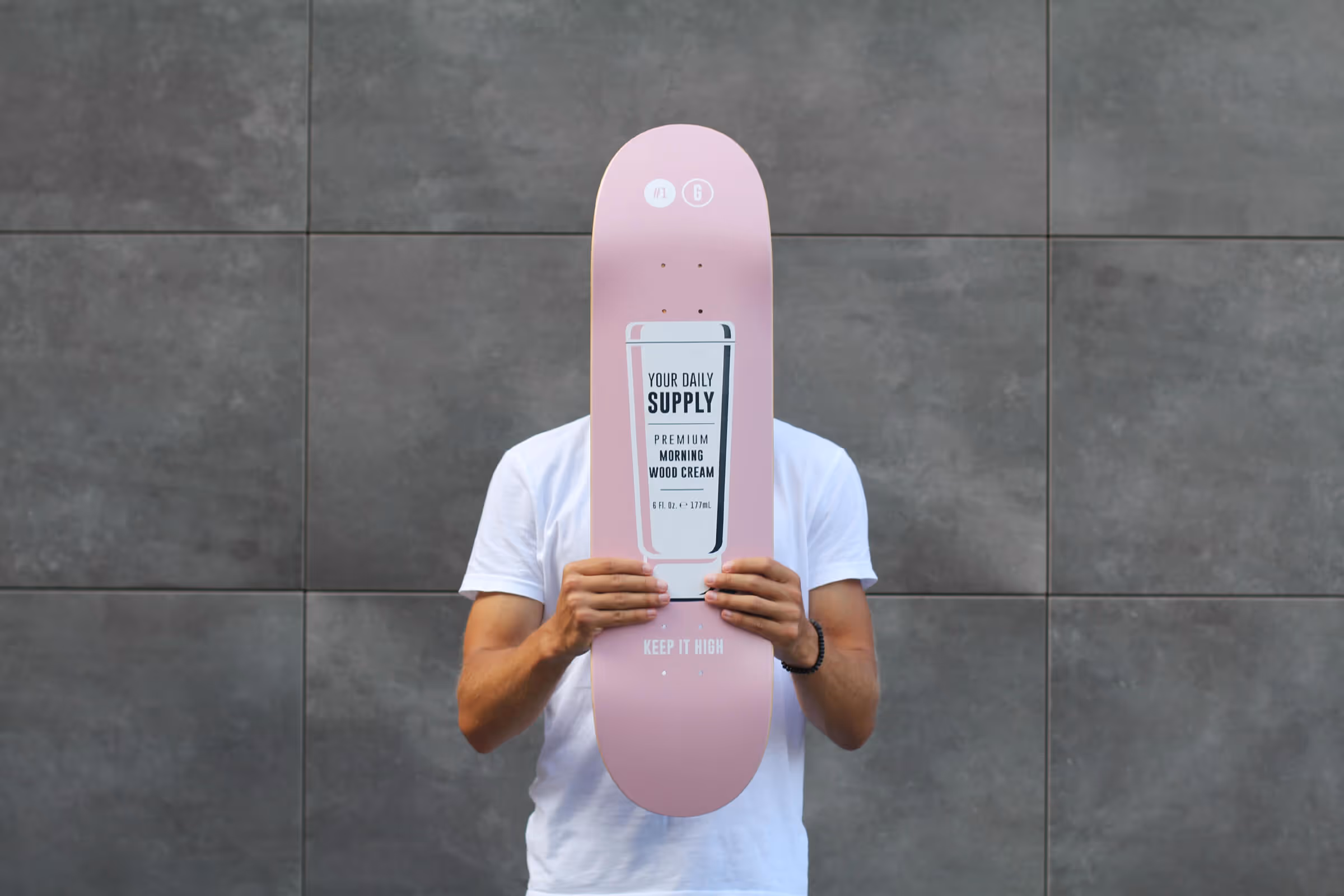 Person in a white t-shirt holding a pink skateboard with a design that reads 'Your Daily Supply Premium Morning Wood Cream' in front of their face.