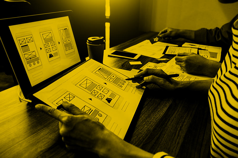People working on wireframe designs for a website on a tablet and printed sheets at a wooden desk.