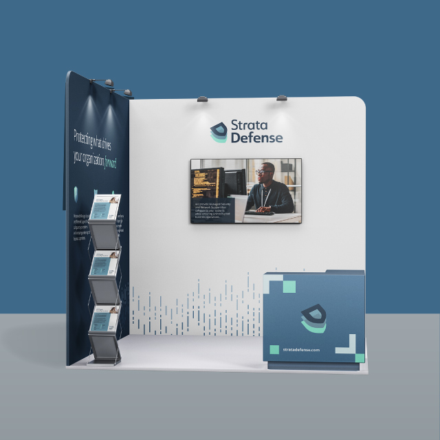 Strata Defense trade show booth with a white and navy blue backdrop, company logo, digital screen showing a man working on a computer, brochure stand, and branded counter.