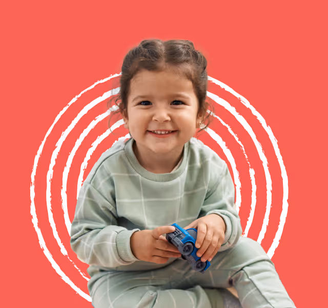 Smiling young child in green pajamas holding a blue toy car, sitting against a red background with white concentric circles.