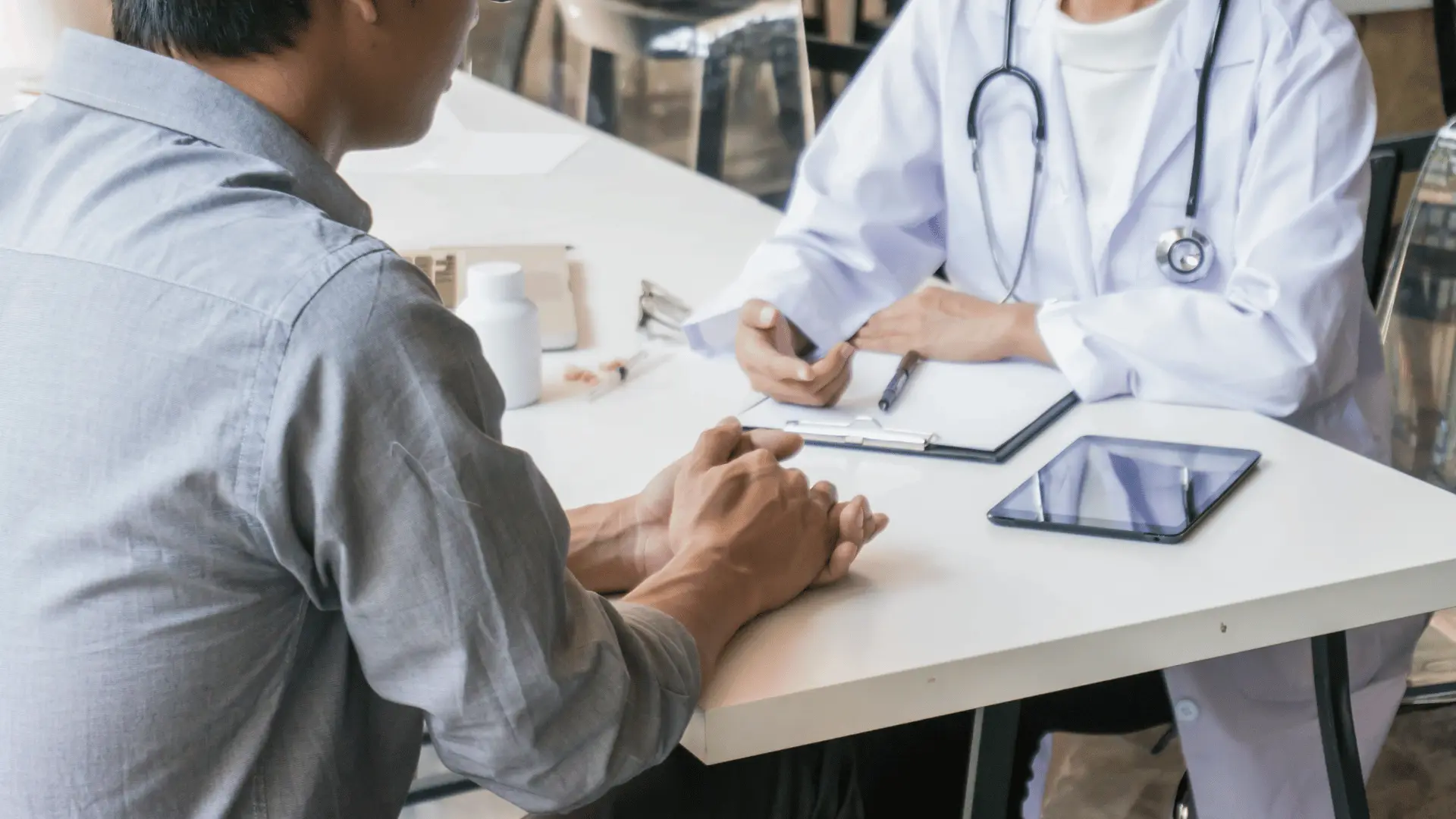 Ein Patient im Beratungsgespräch mit einer Ärztin an einem weißen Schreibtisch, auf dem ein Tablet und medizinische Unterlagen liegen.