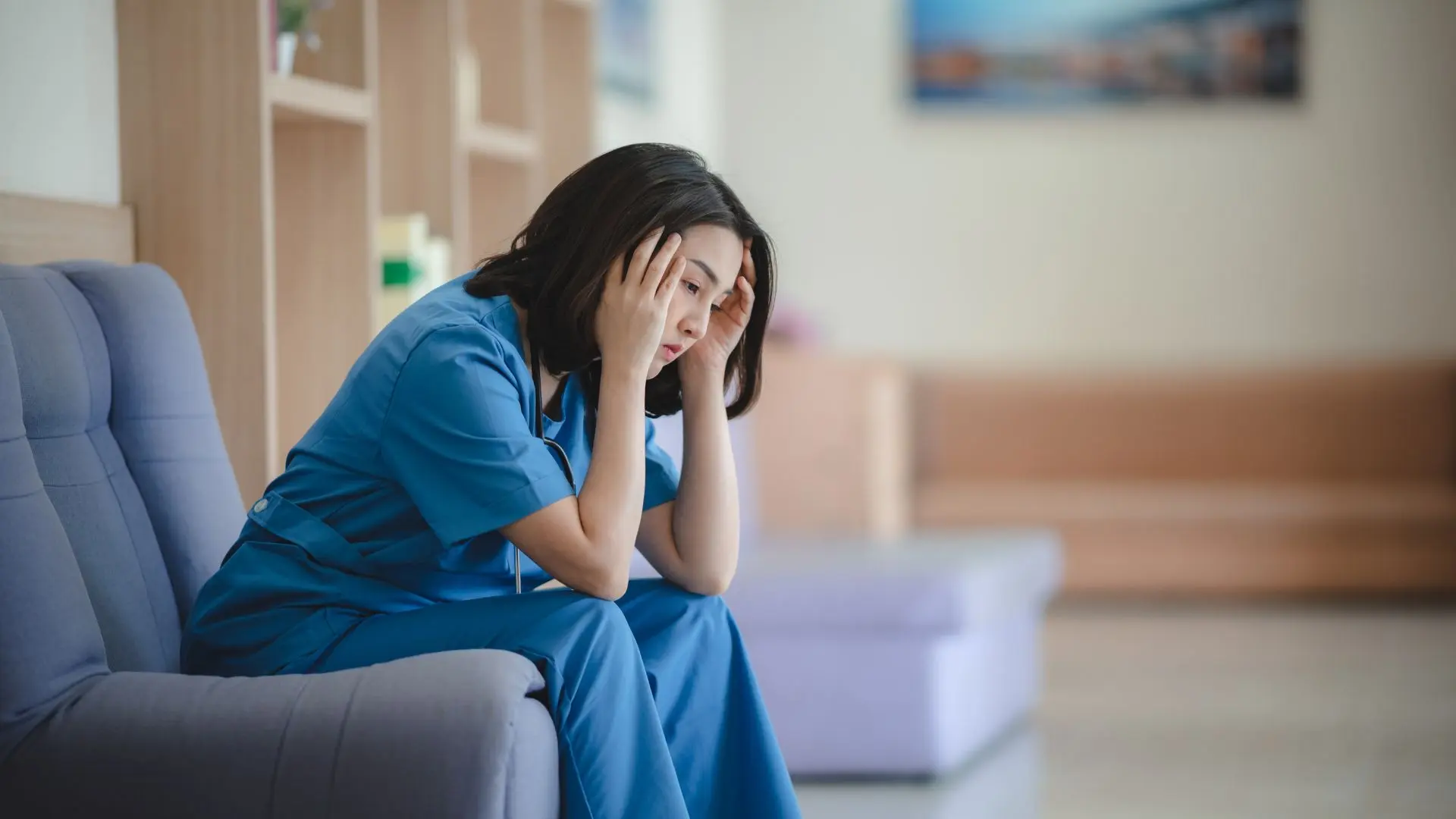 A close-up shows a young Asian woman in blue protective clothing (a nurse or doctor) sitting visibly exhausted and stressed on a sofa. She holds her head in both hands, expressing burnout, emotional exhaustion, and the enormous strain on healthcare professionals. The background is a blurred waiting room or hospital lobby. The image symbolizes the staffing crisis.