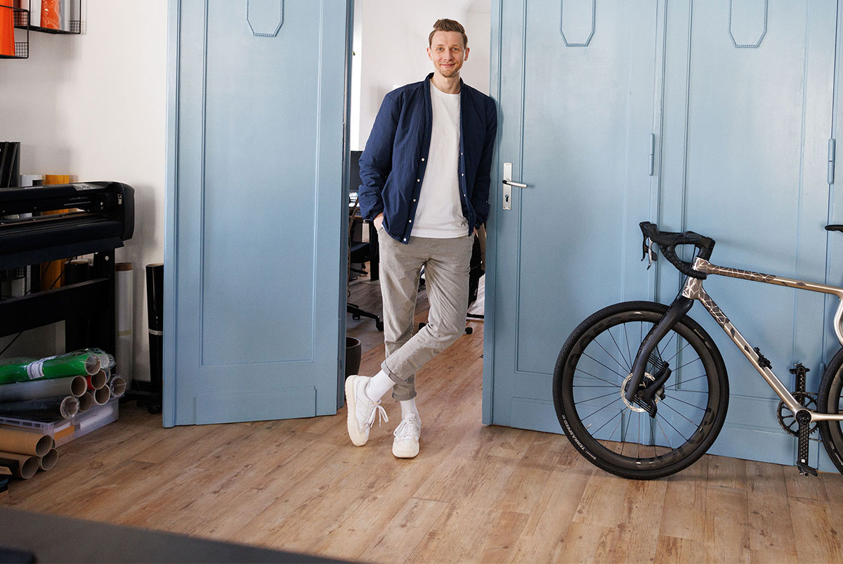 Ein junger Mann mit kurzer Frisur und leichtem Bart steht entspannt in einer offenen Tür mit hellblauen Holztüren. Er trägt eine dunkelblaue Jacke, ein weißes Shirt, graue Hosen und weiße Sneakers. Sein linkes Bein ist locker vor das rechte gestellt. Rechts im Bild lehnt ein modernes Rennrad mit Carbonrahmen an der Wand. Links sind Drucker, Papierrollen und weiteres Büromaterial zu sehen, was auf ein kreatives Arbeitsumfeld hindeutet.
